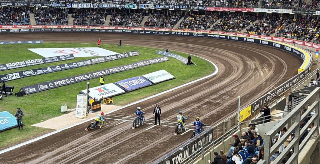 Speedway Race Start Four Riders on Dirt Bikes Lined Up on a Curved Oval Track with Crowd Stands and Advertising Banners Around the Arena 9 Września 2025 
