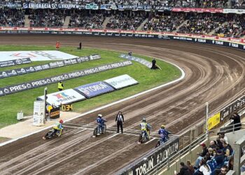 Speedway Race Start Four Riders on Dirt Bikes Lined Up on a Curved Oval Track with Crowd Stands and Advertising Banners Around the Arena 6 Sierpnia 2023 