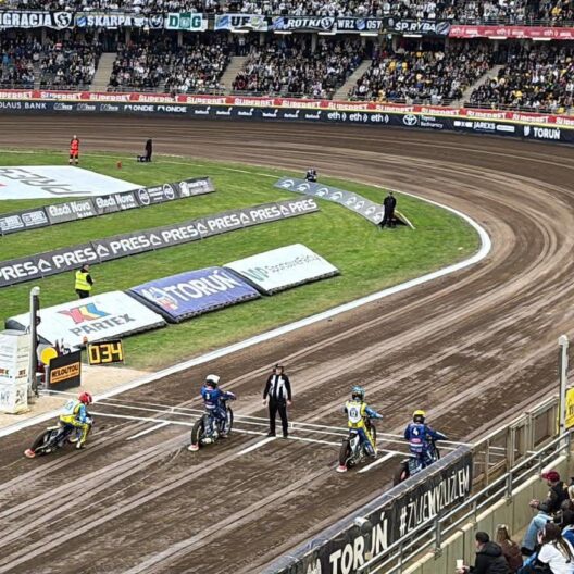 Speedway Race Start Four Riders on Dirt Bikes Lined Up on a Curved Oval Track with Crowd Stands and Advertising Banners Around the Arena 9 Września 2025 