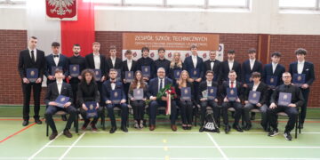 Group of students in suits posing for a formal graduation photo in a school gym, holding blue diplomas.