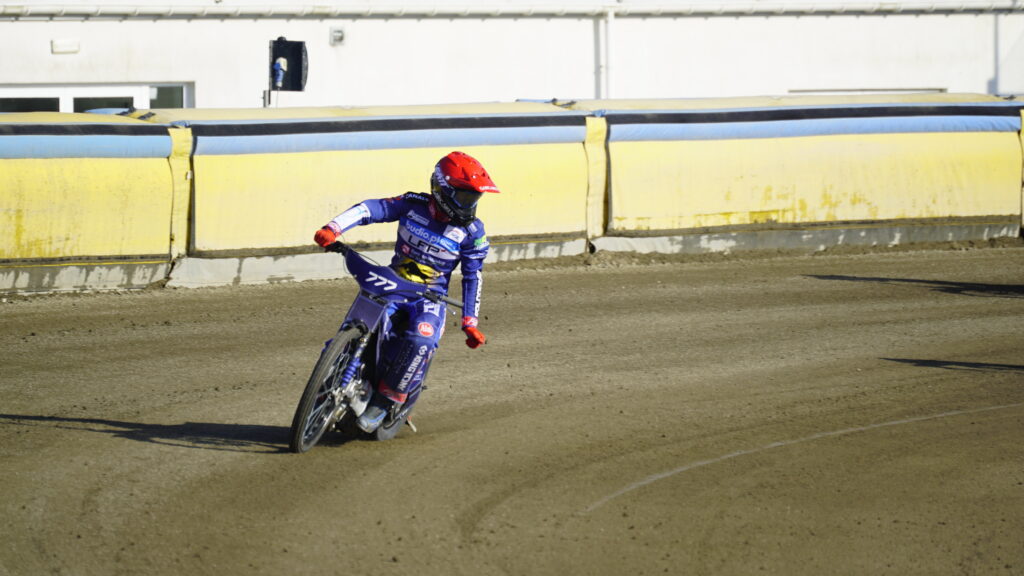 Motocross Rider in Blue Gear and Red Helmet Leaning into a Turn on a Dirt Track Beside Yellow Barriers Behind a Building Edge 9 Września 2025 