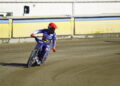 Motocross Rider in Blue Gear and Red Helmet Leaning into a Turn on a Dirt Track Beside Yellow Barriers Behind a Building Edge 15 Czerwca 2020 