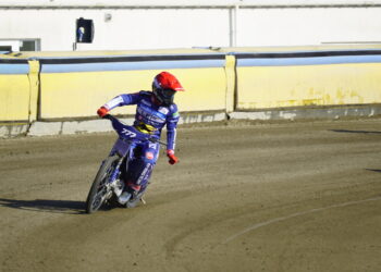 Motocross Rider in Blue Gear and Red Helmet Leaning into a Turn on a Dirt Track Beside Yellow Barriers Behind a Building Edge 19 Kwietnia 2024 