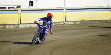 Motocross Rider in Blue Gear and Red Helmet Leaning into a Turn on a Dirt Track Beside Yellow Barriers Behind a Building Edge 30 Czerwca 2024 