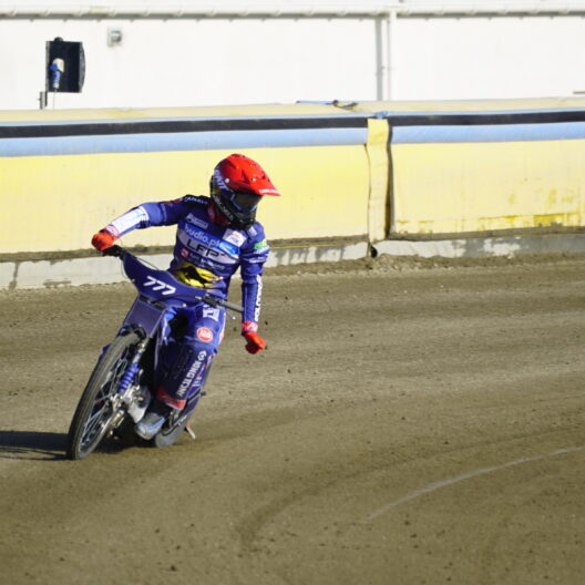 Motocross Rider in Blue Gear and Red Helmet Leaning into a Turn on a Dirt Track Beside Yellow Barriers Behind a Building Edge 9 Września 2025 
