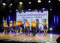 Kids Performing a Dance on Stage in Yellow Shirts and Blue Bottoms with a Banner Reading v MiĘdzyprzedszkolny Festiwal TaŃca in the Background 16 Czerwca 2020  ml 2358