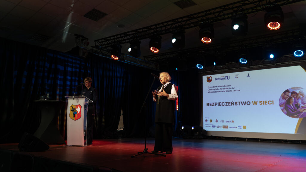 Older Man in a Vest Speaks at a Podium on a Lit Stage with a Projection Screen Behind Him Showing a Presentation and Crest on the Podium 9 Września 2025  ml 2602