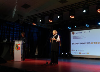 Older Man in a Vest Speaks at a Podium on a Lit Stage with a Projection Screen Behind Him Showing a Presentation and Crest on the Podium 28 Października 2022  ml 2602