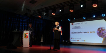 Older Man in a Vest Speaks at a Podium on a Lit Stage with a Projection Screen Behind Him Showing a Presentation and Crest on the Podium 16 Czerwca 2020  ml 2602