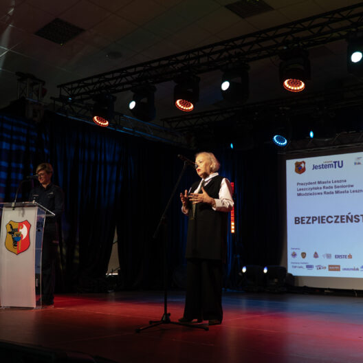 Older Man in a Vest Speaks at a Podium on a Lit Stage with a Projection Screen Behind Him Showing a Presentation and Crest on the Podium 9 Września 2025  ml 2602