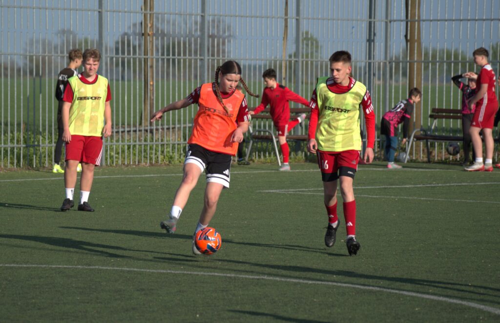 Youth Soccer Scene Player in an Orange Bib Dribbles an Orange Ball While Teammates in Neon Bibs Stand Nearby Near a Tall Fence 9 Września 2025 