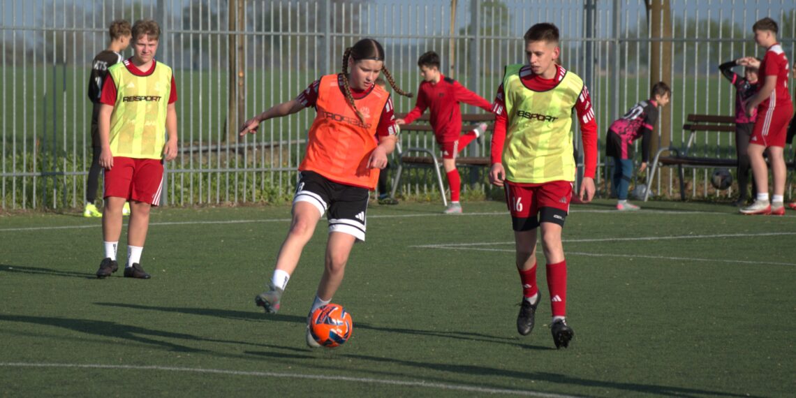 Youth Soccer Scene Player in an Orange Bib Dribbles an Orange Ball While Teammates in Neon Bibs Stand Nearby Near a Tall Fence 30 Czerwca 2024 