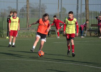 Youth Soccer Scene Player in an Orange Bib Dribbles an Orange Ball While Teammates in Neon Bibs Stand Nearby Near a Tall Fence 8 Kwietnia 2024 