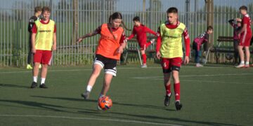 Youth Soccer Scene Player in an Orange Bib Dribbles an Orange Ball While Teammates in Neon Bibs Stand Nearby Near a Tall Fence 14 Czerwca 2020