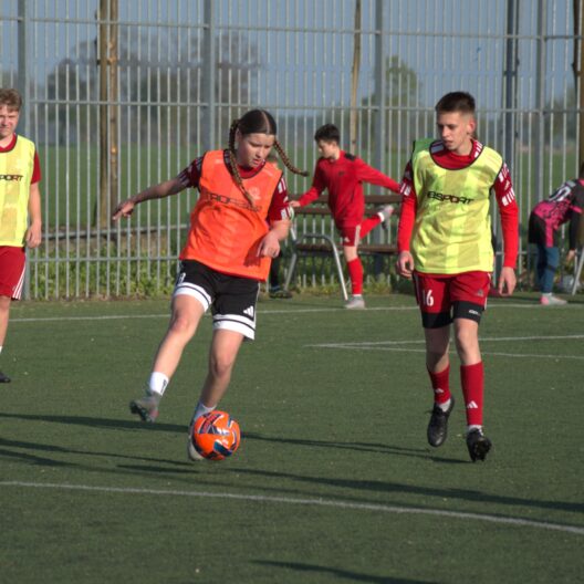 Youth Soccer Scene Player in an Orange Bib Dribbles an Orange Ball While Teammates in Neon Bibs Stand Nearby Near a Tall Fence 9 Września 2025 