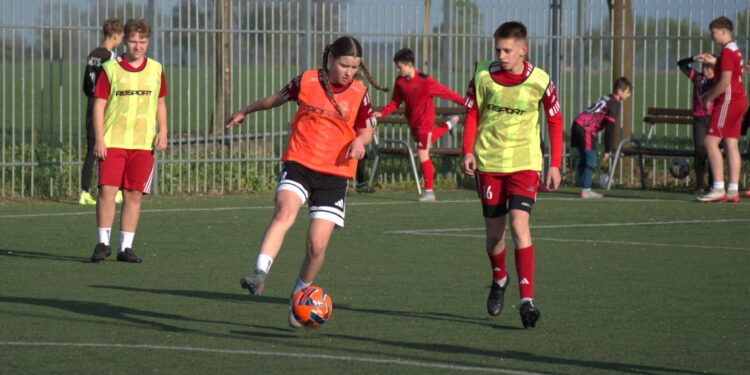 Youth Soccer Scene Player in an Orange Bib Dribbles an Orange Ball While Teammates in Neon Bibs Stand Nearby Near a Tall Fence 26 Kwietnia 2026 