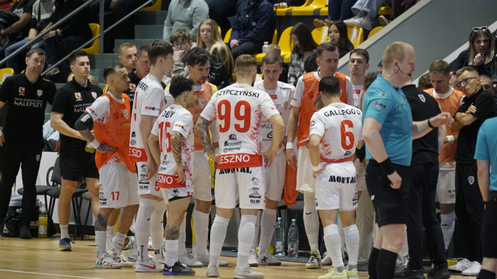Handball Team in White and Red Uniforms Huddles on the Court Orange Bibs Visible Spectators in Yellow Seats Behind Them 9 Września 2025 