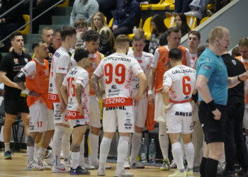 Handball Team in White and Red Uniforms Huddles on the Court Orange Bibs Visible Spectators in Yellow Seats Behind Them 27 Października 2024 