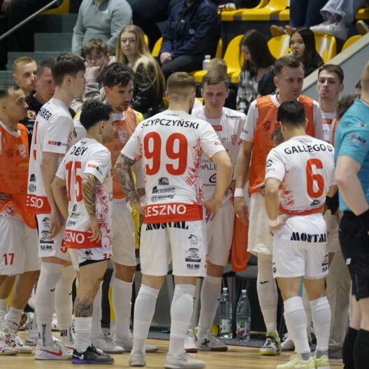 Handball Team in White and Red Uniforms Huddles on the Court Orange Bibs Visible Spectators in Yellow Seats Behind Them 9 Września 2025 