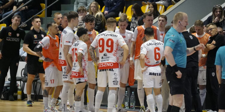 Handball Team in White and Red Uniforms Huddles on the Court Orange Bibs Visible Spectators in Yellow Seats Behind Them 6 Maja 2026 
