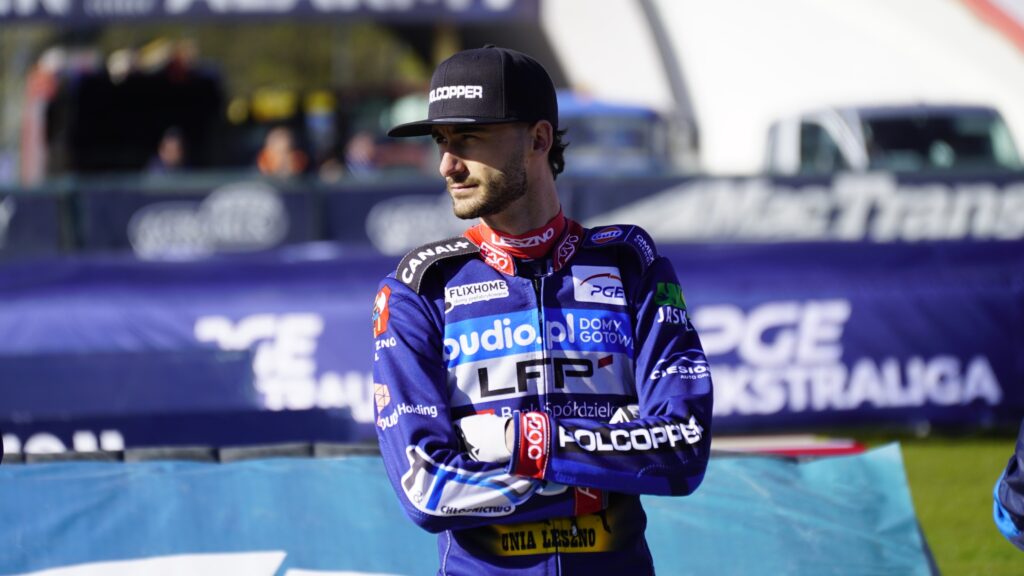 Male Motorcycle Racer in Blue Racing Suit and Black Cap Standing with Arms Crossed at a Track Blurred Banners in the Background 9 Września 2025  ml 3425