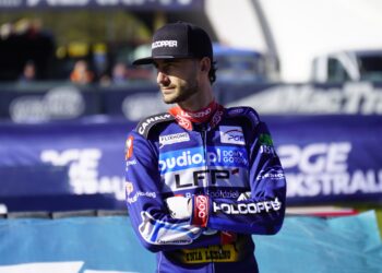 Male Motorcycle Racer in Blue Racing Suit and Black Cap Standing with Arms Crossed at a Track Blurred Banners in the Background 27 Kwietnia 2023  ml 3425