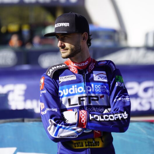 Male Motorcycle Racer in Blue Racing Suit and Black Cap Standing with Arms Crossed at a Track Blurred Banners in the Background 9 Września 2025  ml 3425