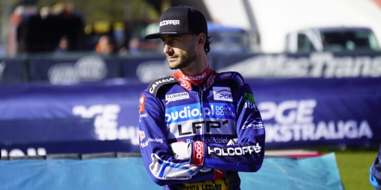 Male Motorcycle Racer in Blue Racing Suit and Black Cap Standing with Arms Crossed at a Track Blurred Banners in the Background 25 Maja 2026  ml 3425