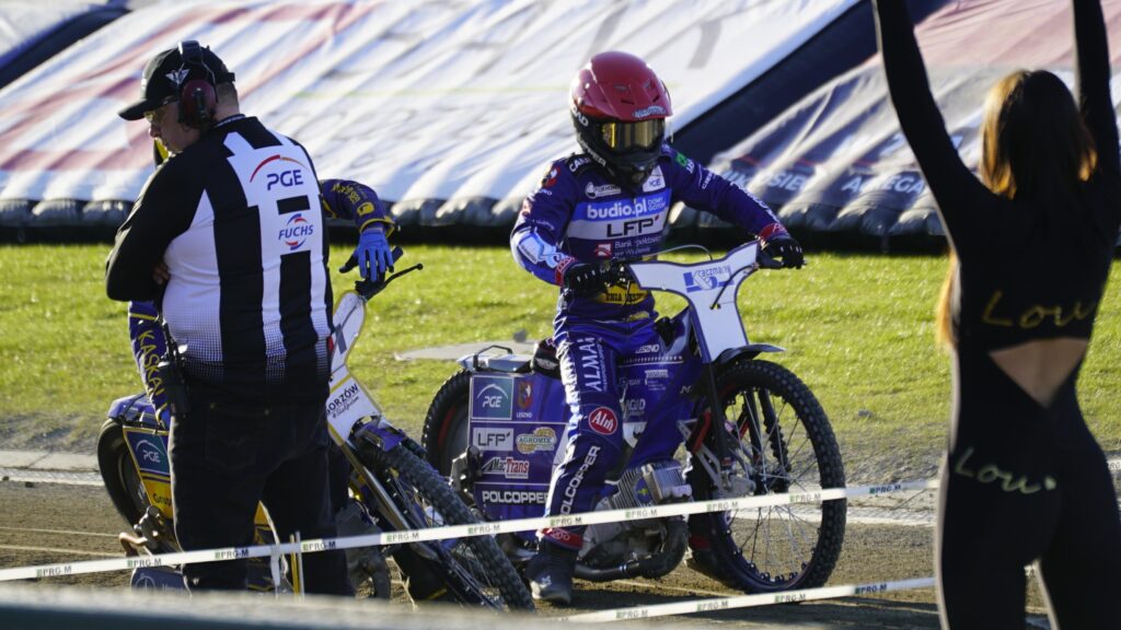 Motocross Race Scene with a Rider in Purple Gear and Red Helmet on a Blue Bike Paused Near the Track While Two People Stand Nearby 9 Września 2025 