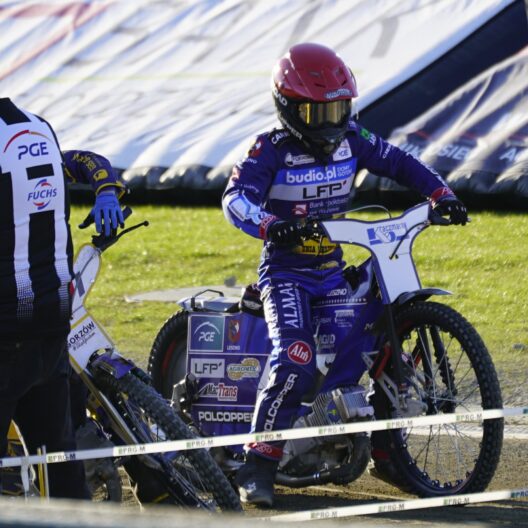 Motocross Race Scene with a Rider in Purple Gear and Red Helmet on a Blue Bike Paused Near the Track While Two People Stand Nearby 9 Września 2025 