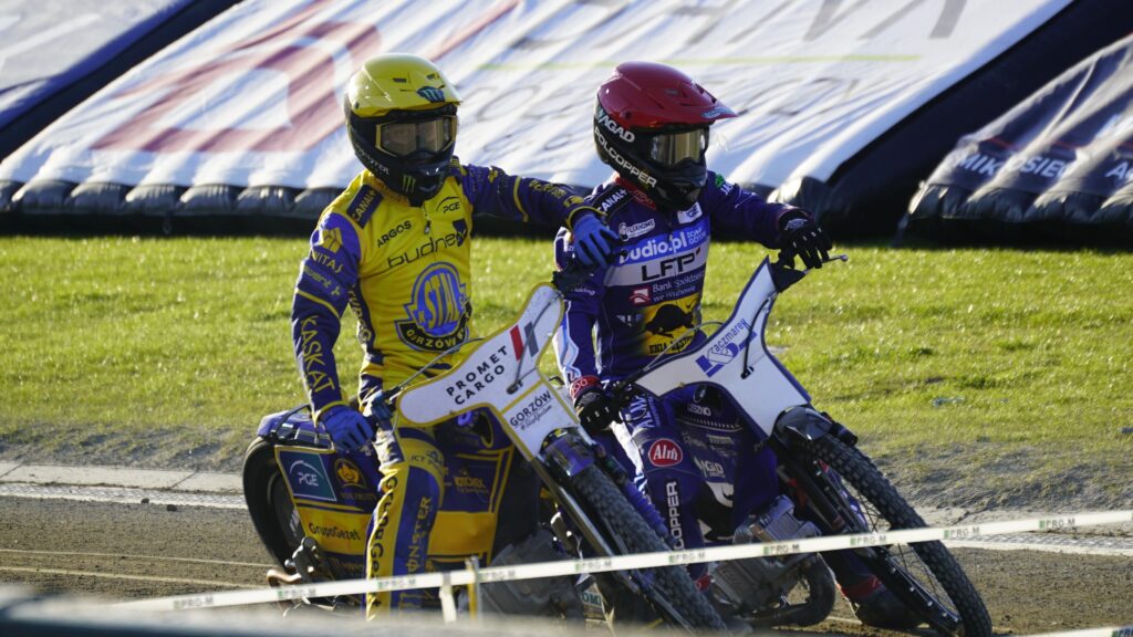 Two Speedway Riders in Yellow and Purple Racing Suits Lean into a Turn on a Dirt Track with Grass in the Background 9 Września 2025 