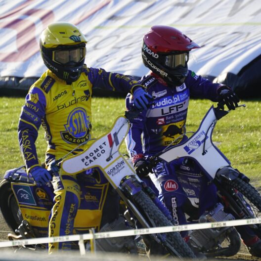Two Speedway Riders in Yellow and Purple Racing Suits Lean into a Turn on a Dirt Track with Grass in the Background 9 Września 2025 