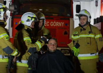 Firefighters in Tan Turnout Gear and Helmets Surround a Seated Man with a Red Fire Engine in the Background Inside a Station 1 Czerwca 2023 
