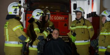 Firefighters in tan turnout gear and helmets surround a seated man, with a red fire engine in the background inside a station.