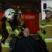 Firefighters in Tan Turnout Gear and Helmets Surround a Seated Man with a Red Fire Engine in the Background Inside a Station 15 Czerwca 2020 