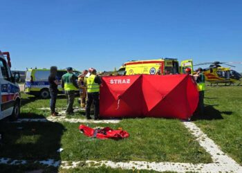 Emergency Responders Set Up a Bright Red Tent Labeled straŻ on a Grassy Field with Ambulances and a Yellow Helicopter in the Background 15 Sierpnia 2021 