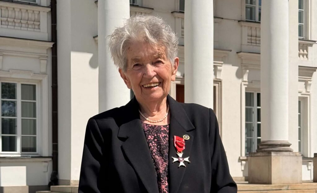 Smiling Elderly Woman in a Black Blazer with a Red Ribbon Medal Standing Outside a White Columned Building 9 Września 2025