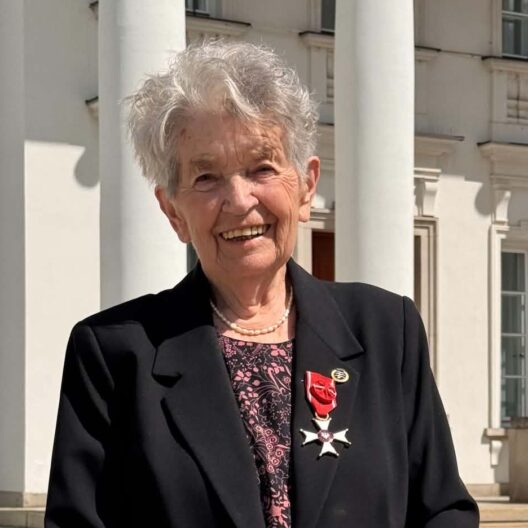 Smiling Elderly Woman in a Black Blazer with a Red Ribbon Medal Standing Outside a White Columned Building 9 Września 2025