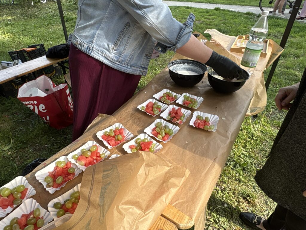 Person in a Denim Jacket Arranges Fruit Skewers of Watermelon and Green Grapes on Paper Liners at an Outdoor Picnic Table in a Park Park Setting 9 Września 2025