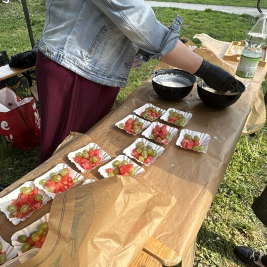Person in a Denim Jacket Arranges Fruit Skewers of Watermelon and Green Grapes on Paper Liners at an Outdoor Picnic Table in a Park Park Setting 9 Września 2025
