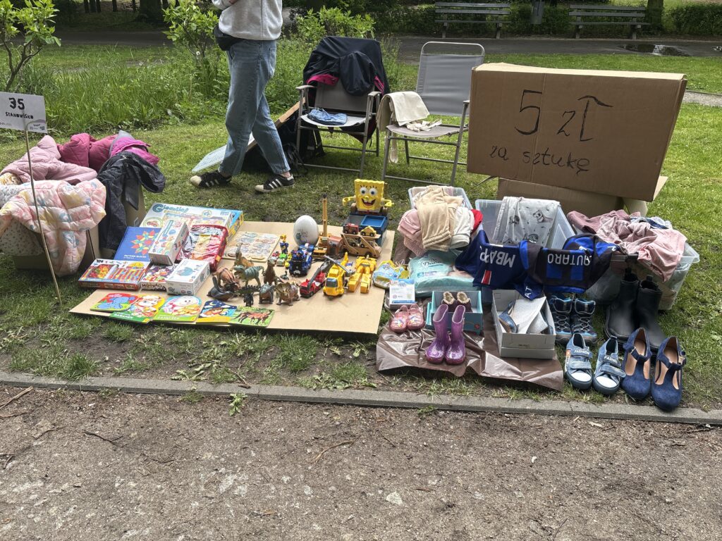 Outdoor Yard Sale Setup Blankets and Boxes on Grass Display Clothing Blankets Toys and Shoes with a Handwritten Price Sign Nearby 9 Września 2025