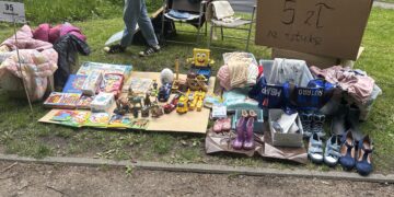 Outdoor yard sale setup: blankets and boxes on grass display clothing, blankets, toys, and shoes with a handwritten price sign nearby.