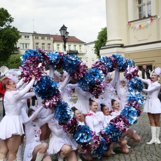 Cheerleaders in White Dresses Performing with Blue and Pink Pom poms in a Street Parade Band in Red Uniforms and Spectators in the Background 9 Września 2025