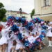 Cheerleaders in White Dresses Performing with Blue and Pink Pom poms in a Street Parade Band in Red Uniforms and Spectators in the Background 15 Czerwca 2020