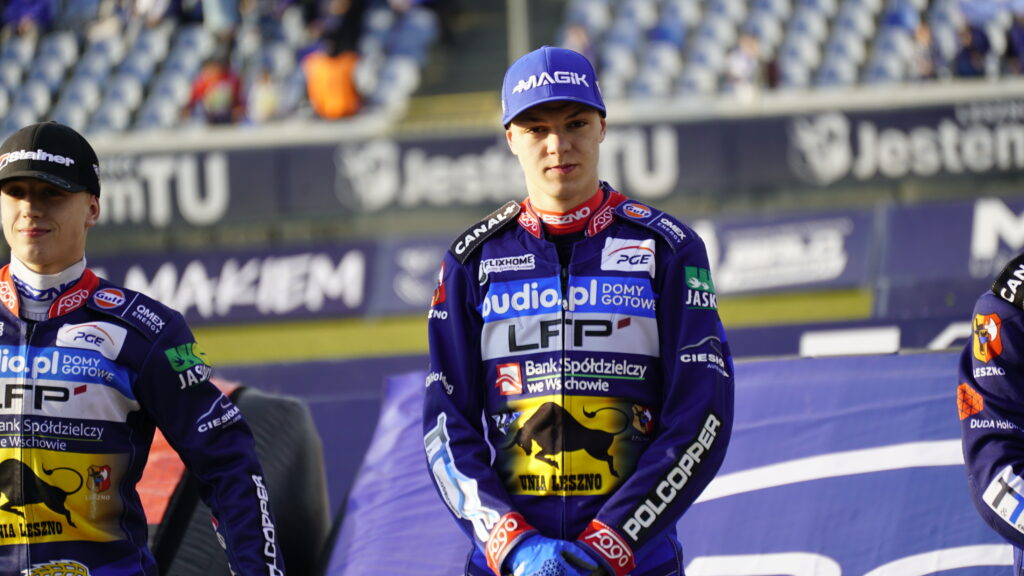 Three Motorcycle Racers in Purple Racing Suits Standing on a Podium with Sponsor Logos Citytrack Backdrop and Blurred Spectators Behind Them Center Rider Looking Ahead Wearing a Blue Cap 9 Września 2025