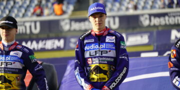 Three Motorcycle Racers in Purple Racing Suits Standing on a Podium with Sponsor Logos Citytrack Backdrop and Blurred Spectators Behind Them Center Rider Looking Ahead Wearing a Blue Cap 12 Czerwca 2020