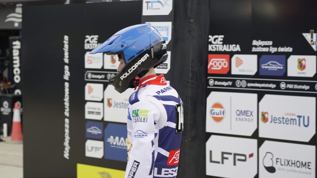 Motorsport Rider in a Blue Helmet and White Racing Suit Stands at a Sponsor filled Pit Wall 9 Września 2025  ml 5067