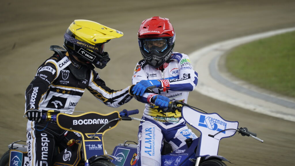 Two Speedway Riders in Colorful Racing Suits on a Dirt Track Shaking Hands Mid race or After a Heat Their Bikes Visible in the Foreground 9 Września 2025  ml 5210