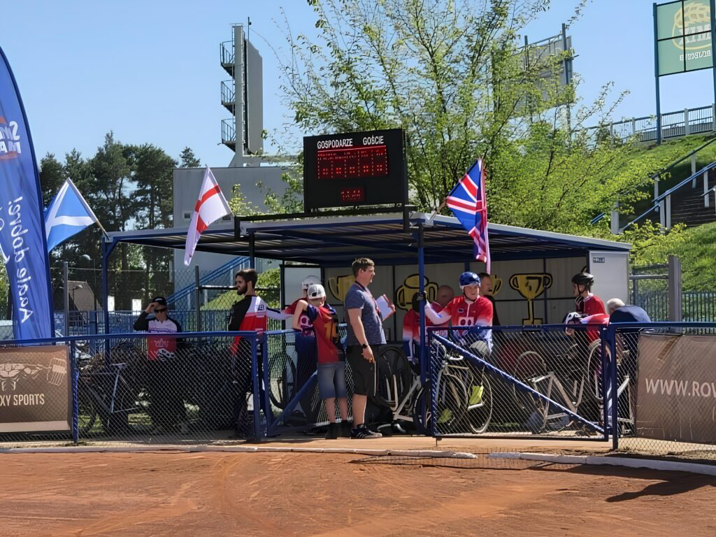 Group of Athletes in Red Uniforms Standing Behind Blue Fencing at an Outdoor Sports Event with a Digital Scoreboard and Flags in the Background 9 Września 2025 
