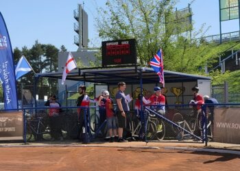 Group of Athletes in Red Uniforms Standing Behind Blue Fencing at an Outdoor Sports Event with a Digital Scoreboard and Flags in the Background 7 Czerwca 2024 
